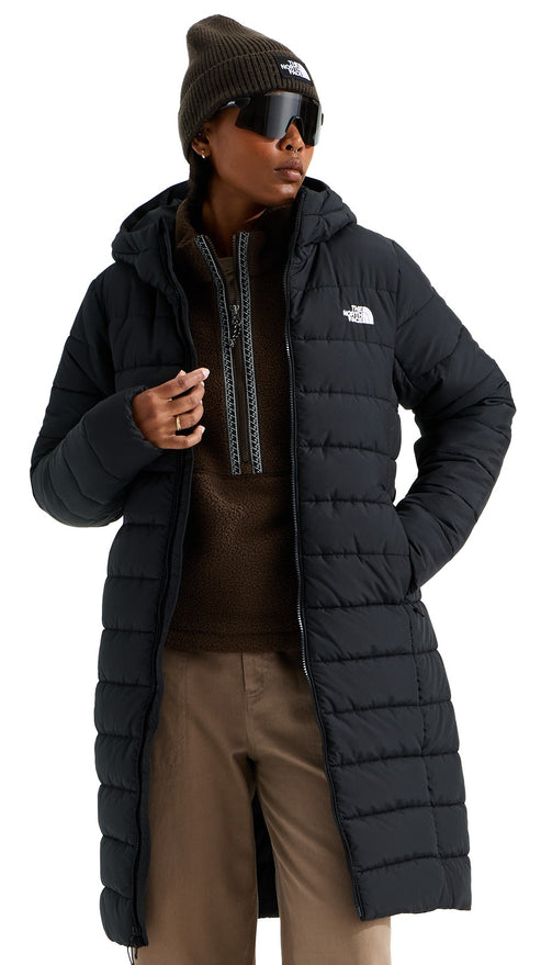 A person wearing a black, knee-length, hooded, quilted puffer coat with a zip-up brown fleece sweater underneath, tan pants, dark sunglasses, and a brown beanie.