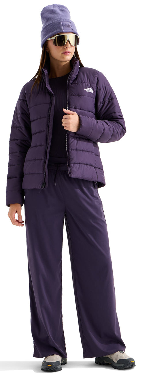 A woman stands wearing a deep purple puffer jacket over a black shirt, paired with matching deep purple wide-leg trousers. She is also wearing a purple beanie, reflective wrap-around sunglasses, and grey and black outdoor sneakers.