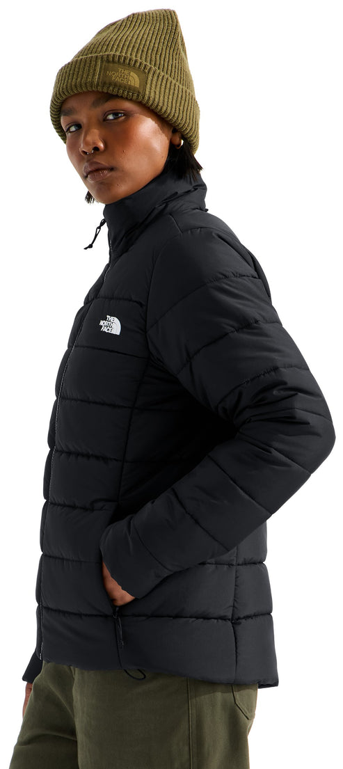 A person in profile wearing a black puffer jacket with horizontal quilting and a white logo on the chest, paired with a green beanie and matching green pants.