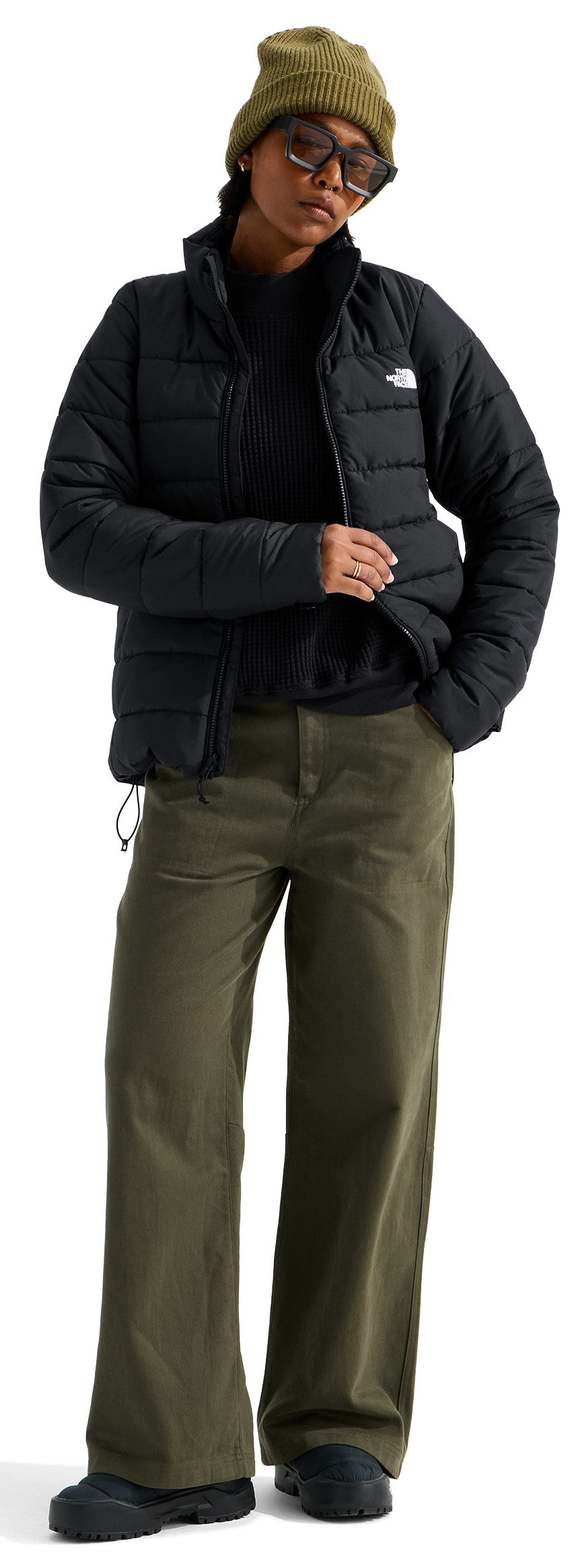 A person posing while wearing a black puffy jacket over a dark sweater, olive green wide-leg trousers, a beige beanie, and dark sunglasses.
