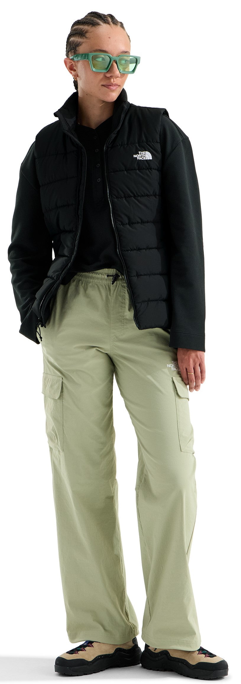 A person with braided hair and green-tinted glasses wearing a black zippered puffer vest over a black shirt, light olive green cargo pants with side pockets, and tan and black casual sneakers.