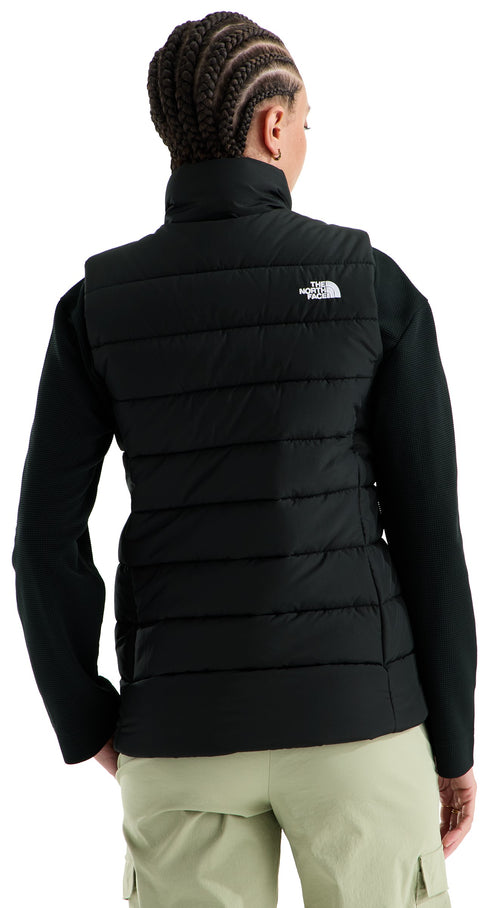 A back view of a person wearing a black quilted puffer vest over a long-sleeved black top and light olive green cargo pants, featuring a small white logo on the upper right back of the vest.