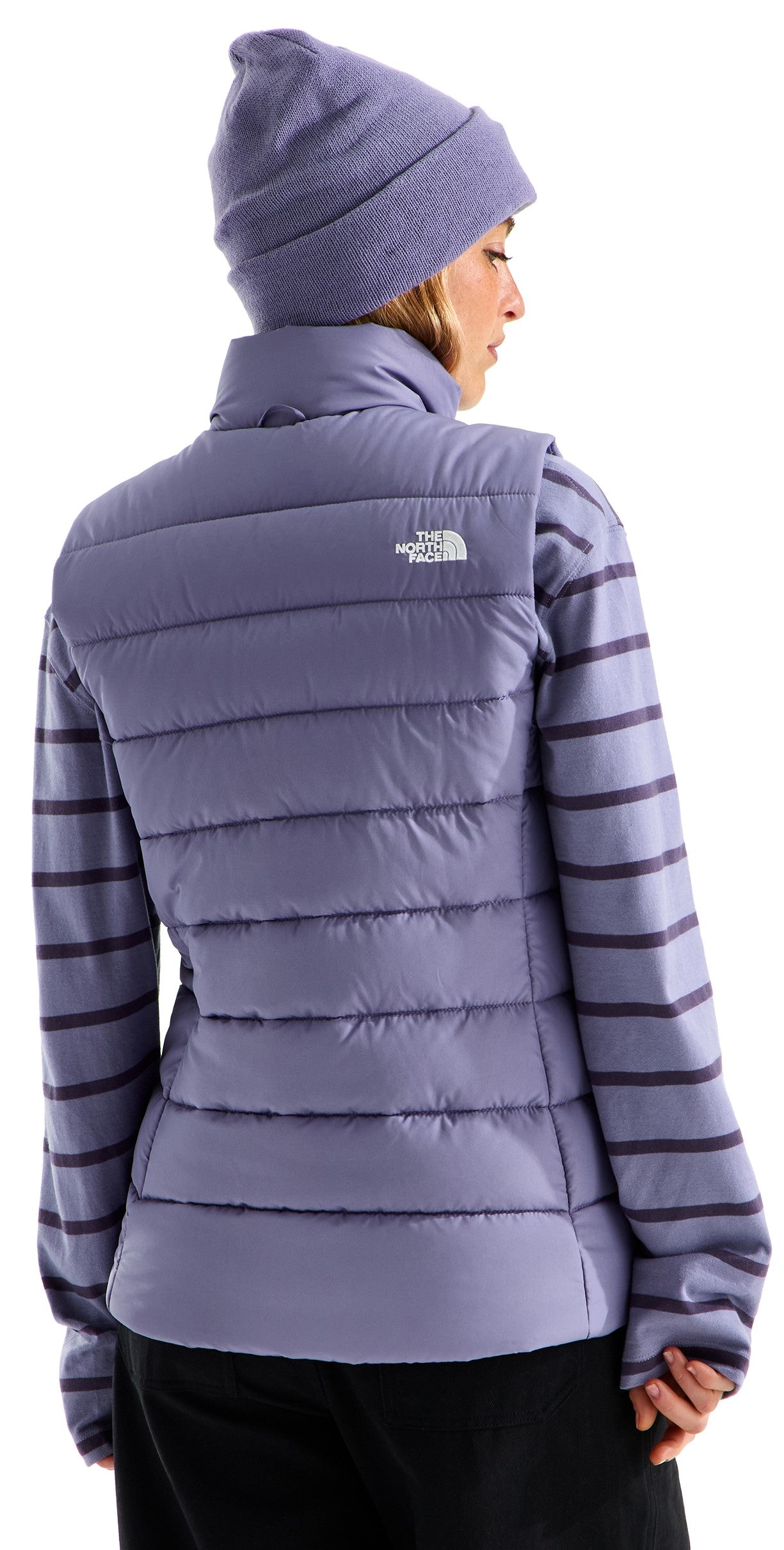 A back view of a person wearing a light purple, horizontal-quilted, sleeveless vest with a mock collar over a matching purple-and-dark-purple horizontal-striped long-sleeved shirt, and a matching light purple knit beanie, against a white background.