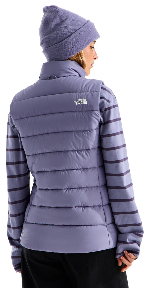 A back view of a person wearing a light purple, horizontal-quilted, sleeveless vest with a mock collar over a matching purple-and-dark-purple horizontal-striped long-sleeved shirt, and a matching light purple knit beanie, against a white background.