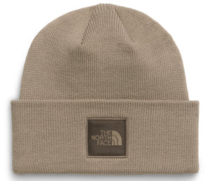 A tan knit cuffed beanie with a square brown patch centered on the cuff featuring the words 'THE NORTH FACE' and a logo icon in a darker brown color.