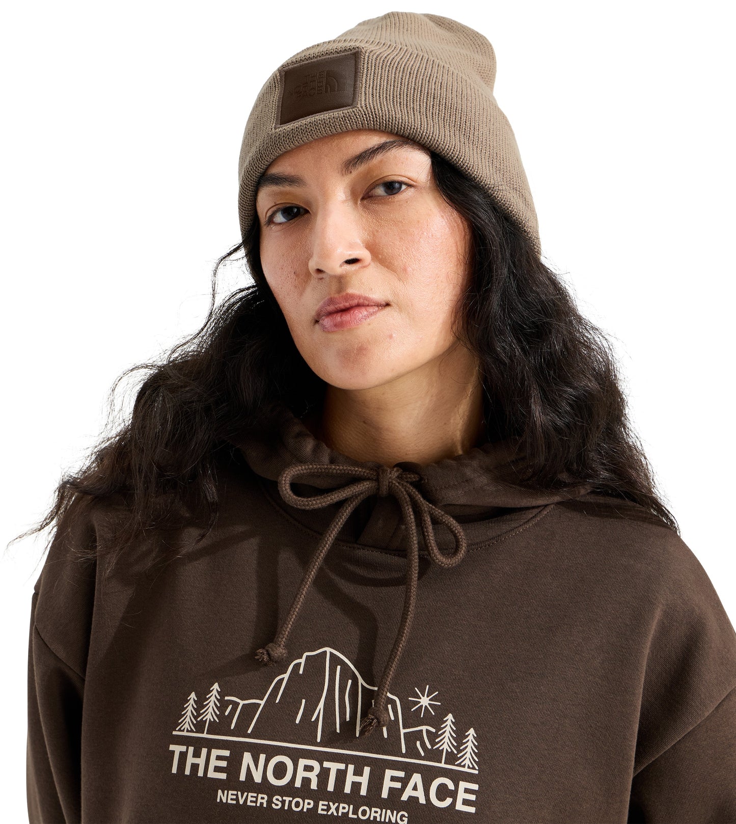 A person with long dark wavy hair wearing a taupe beanie with a rectangular patch and a brown hoodie featuring a white graphic of a mountain, trees, and the text The North Face Never Stop Exploring.