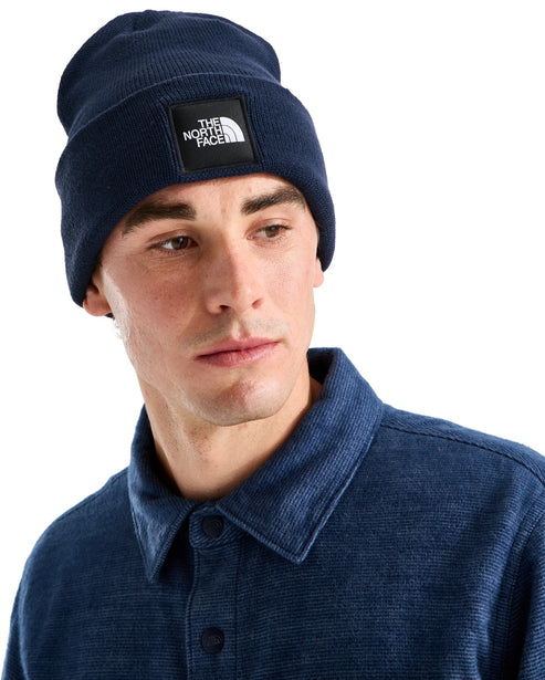 A man wearing a navy blue knit beanie with a square black patch on the front that features a white logo.