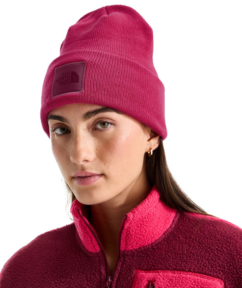 A woman wearing a bright pink knit beanie with a square matching-color patch on the front featuring an embossed circular logo, paired with a two-toned dark and bright pink textured fleece quarter-zip jacket.