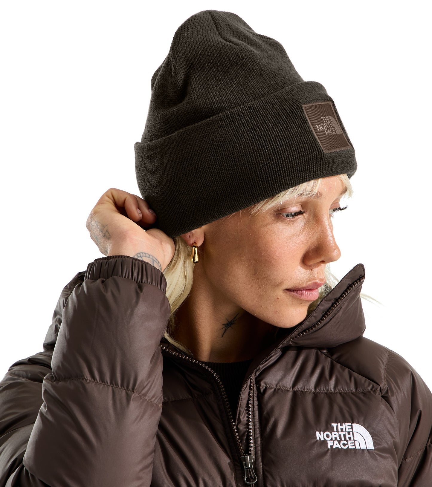 A close-up shot of a person wearing a brown knit beanie with a square brown patch featuring white text, and a matching brown down-filled jacket with a white logo on the chest.