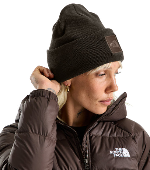 A close-up shot of a person wearing a brown knit beanie with a square brown patch featuring white text, and a matching brown down-filled jacket with a white logo on the chest.