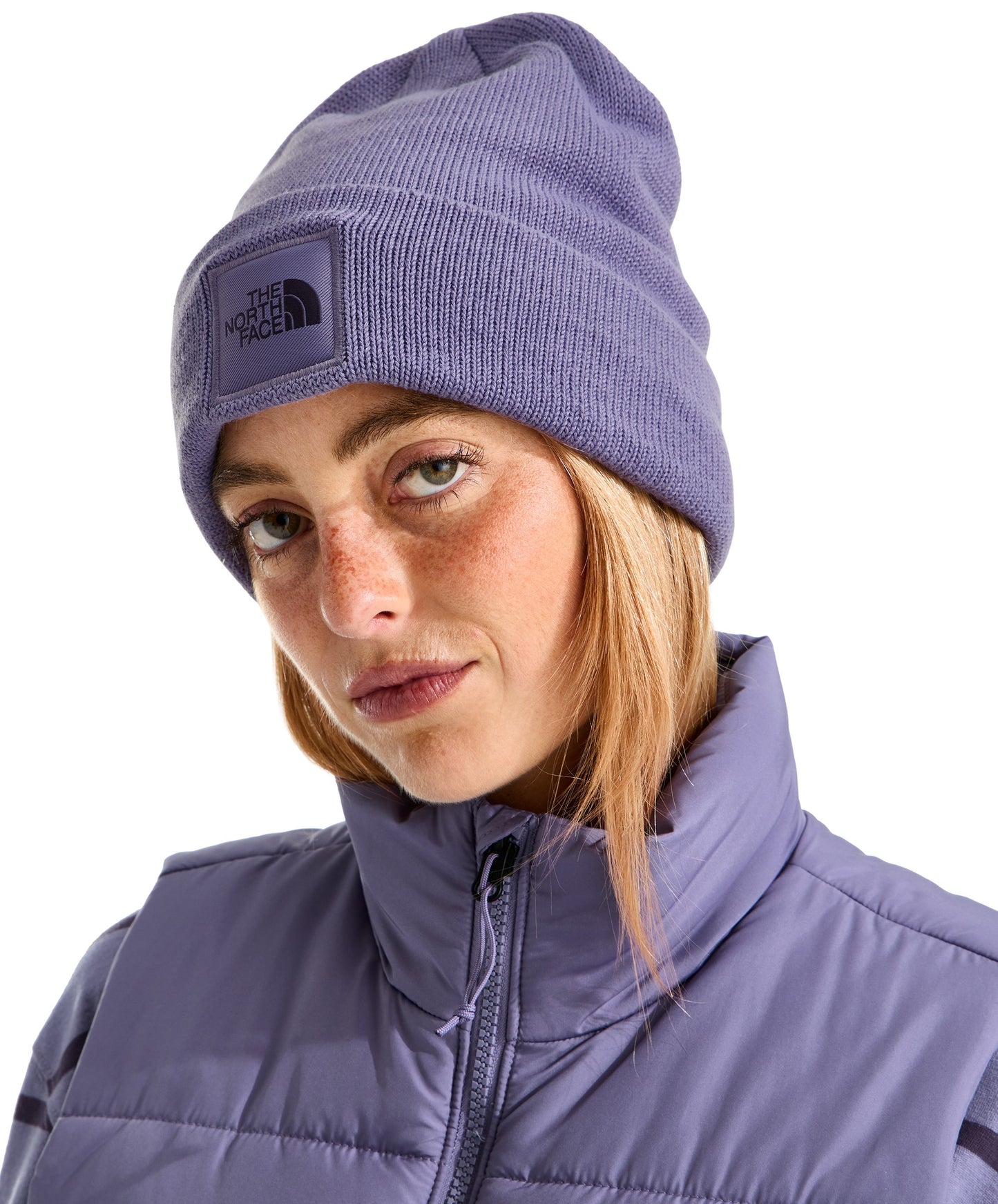 A light-skinned woman with freckles and blonde hair wears a lavender-colored ribbed knit beanie with a square matching patch featuring the words 'THE NORTH FACE' and a mountain silhouette, paired with a matching lavender-colored zip-up quilted vest.