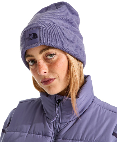 A light-skinned woman with freckles and blonde hair wears a lavender-colored ribbed knit beanie with a square matching patch featuring the words 'THE NORTH FACE' and a mountain silhouette, paired with a matching lavender-colored zip-up quilted vest.