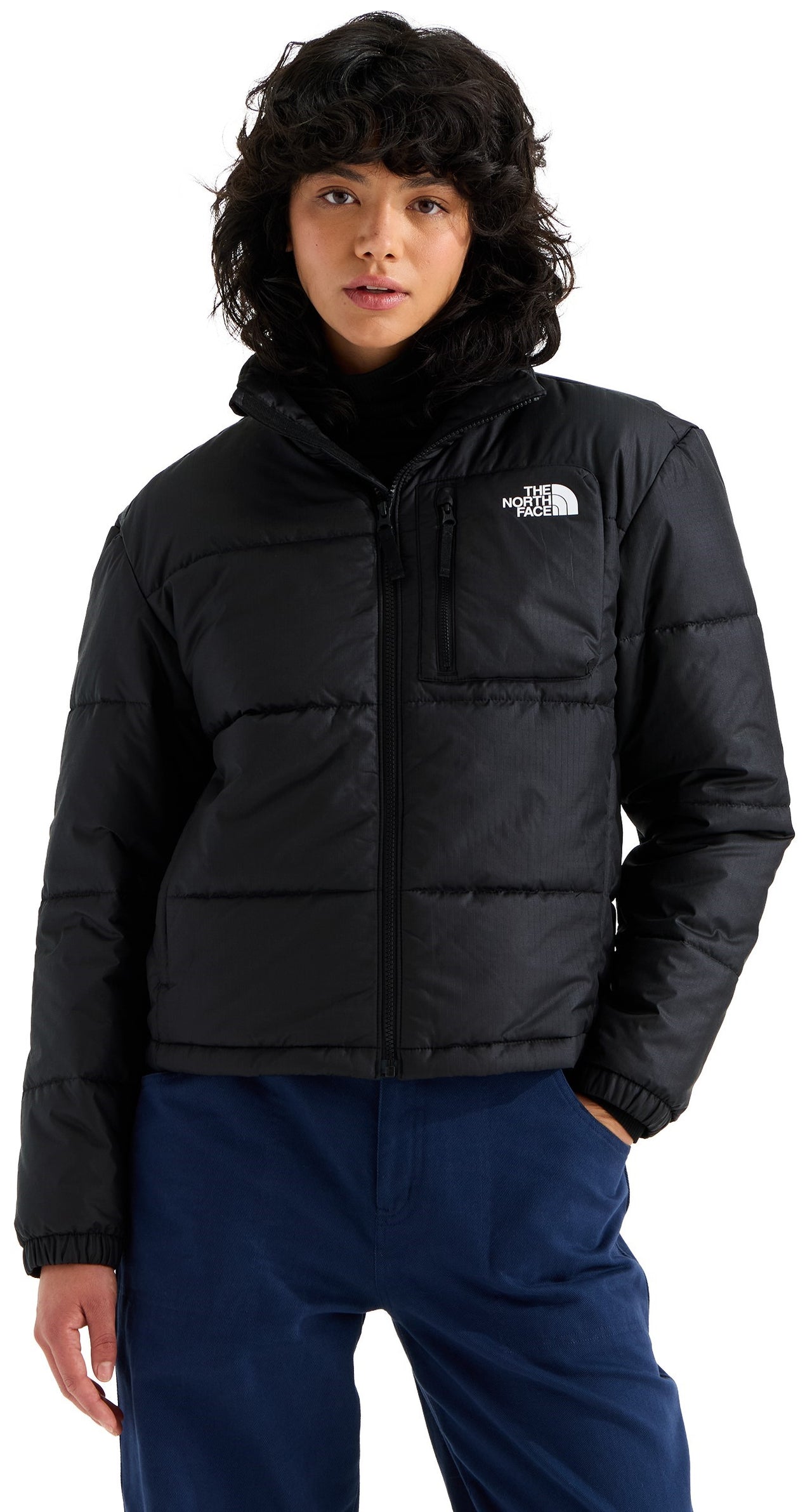 A person with dark, wavy hair wearing a black, zip-up, long-sleeved, high-neck, quilted puffer jacket with a small zippered pocket on the left chest and blue pants.