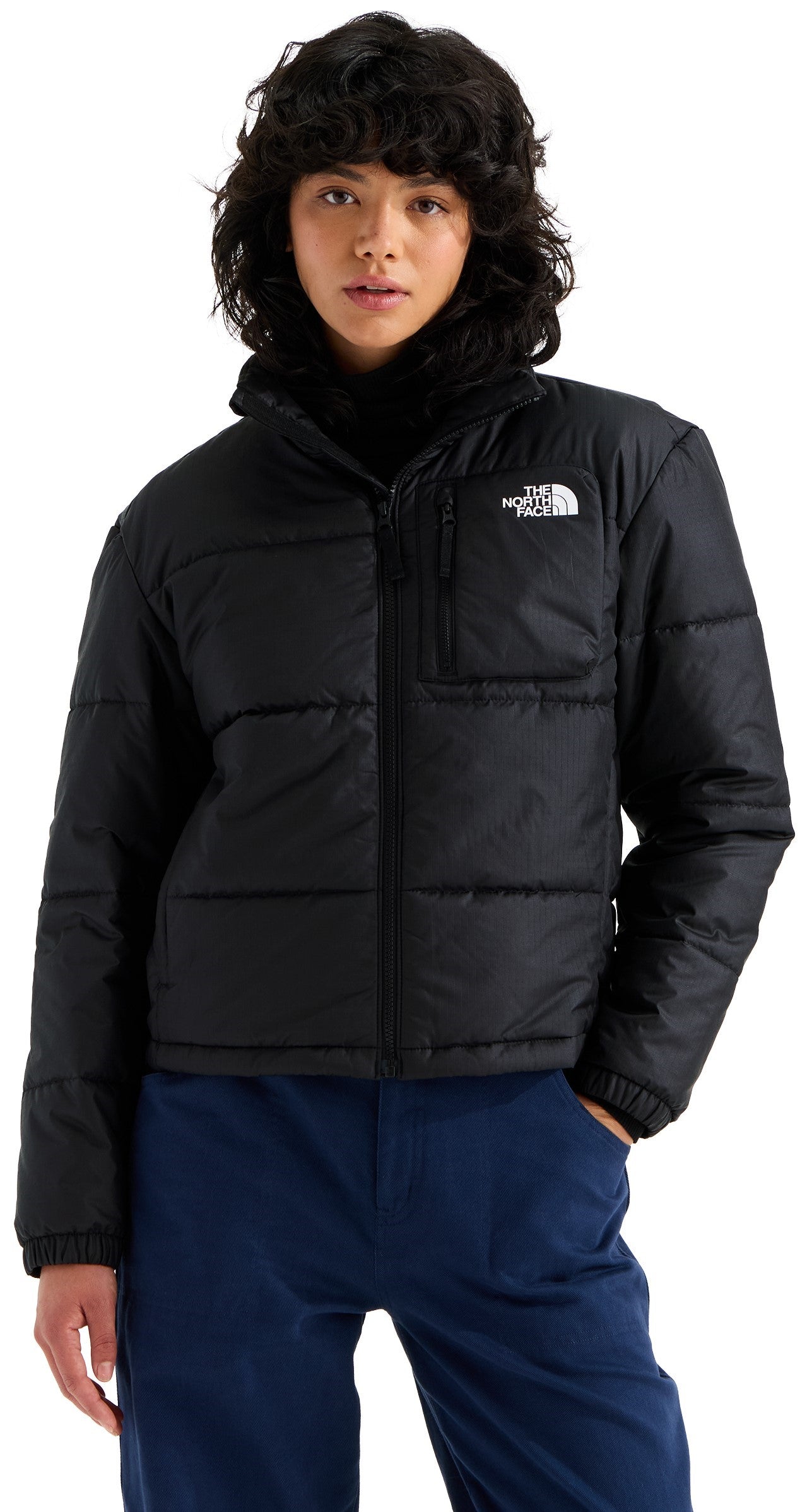 A person with dark, wavy hair wearing a black, zip-up, long-sleeved, high-neck, quilted puffer jacket with a small zippered pocket on the left chest and blue pants.