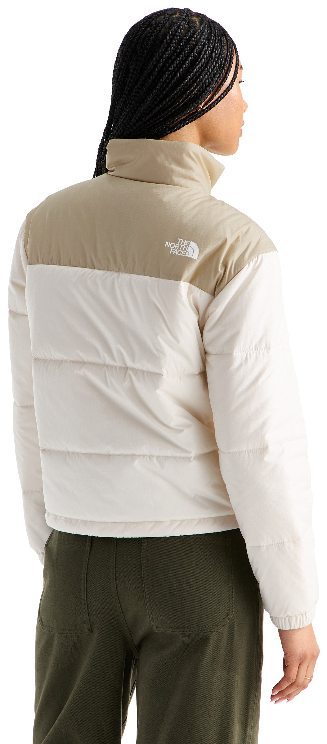 A back view of a person with long braided hair wearing a two-tone, light beige and cream-colored puffer jacket with a high collar and a logo on the upper back, paired with olive green pants.