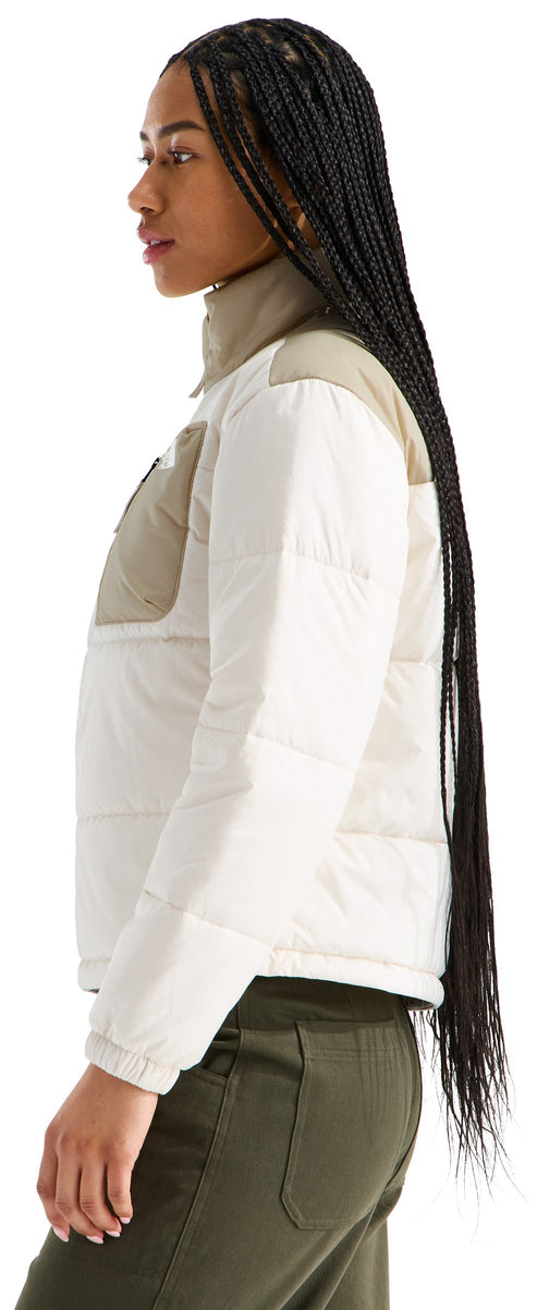 A woman with long black braids seen in profile wears a beige and white color-blocked puffer jacket with a high collar over olive green pants.