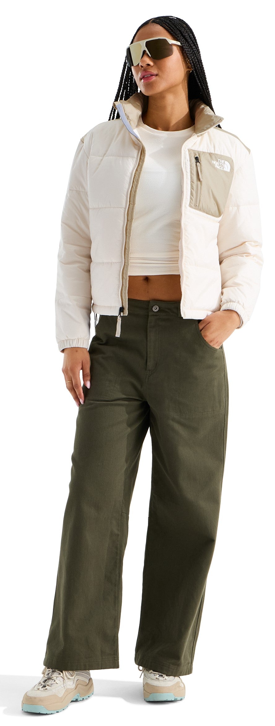 A woman wearing a cropped white jacket with beige accents and a pocket on the left chest, a white crop top, olive green wide-leg pants, and light-colored sneakers, accessorized with tinted sunglasses.