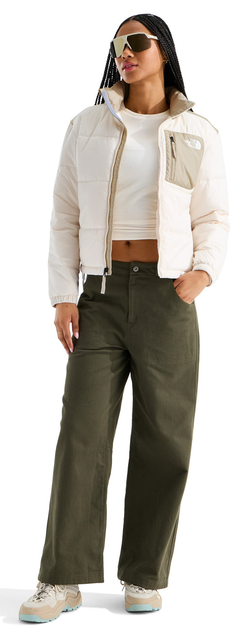 A woman wearing a cropped white jacket with beige accents and a pocket on the left chest, a white crop top, olive green wide-leg pants, and light-colored sneakers, accessorized with tinted sunglasses.