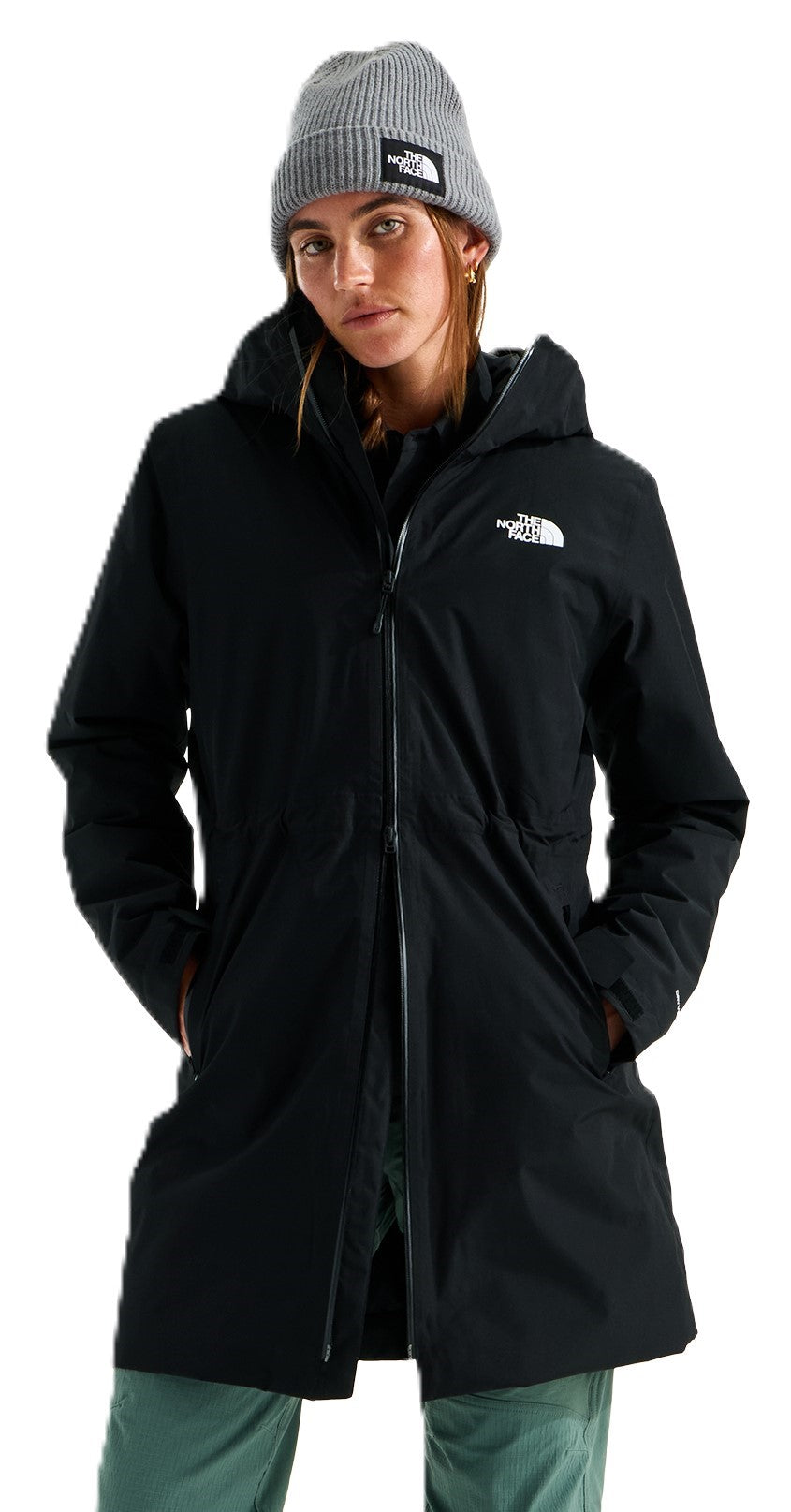 A woman wearing a black, hooded, knee-length, zippered winter coat over a black shirt and green pants, with a gray knit beanie featuring a small patch on the front.