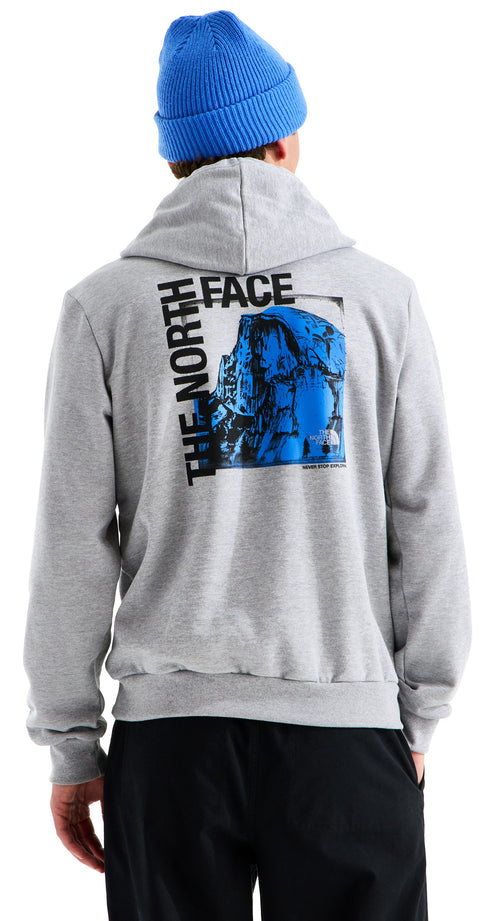 A back view of a person wearing a light gray hooded sweatshirt with a blue beanie. The hoodie features a large square graphic on the back with a blue mountain illustration and the words THE NORTH FACE printed in black vertically along the side.