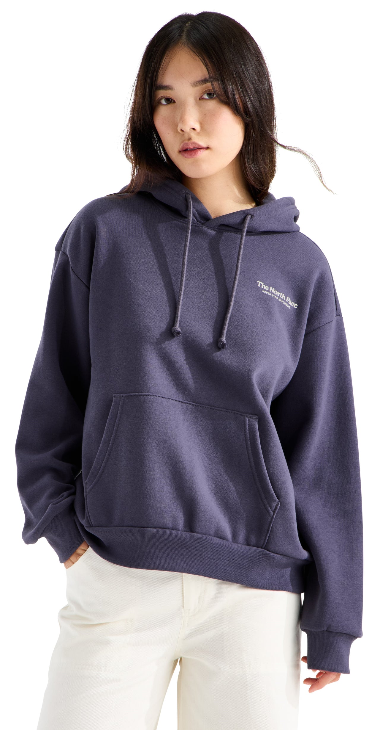 A woman wearing a dark slate blue, long-sleeved, oversized pullover hoodie with a drawstring hood and a front kangaroo pocket, featuring the text "The North Face" in white on the upper left chest, paired with light-colored pants.
