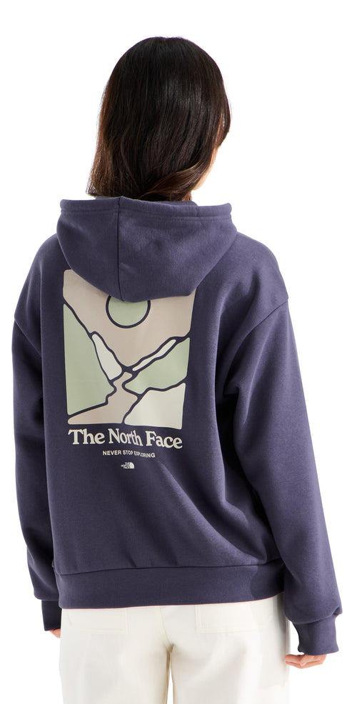 A back view of a person wearing a dark blue hooded sweatshirt featuring a rectangular graphic on the back with an abstract landscape of mountains, a river, and a sun in light green and beige tones, with the text 'The North Face' and 'NEVER STOP EXPLORING' printed below the graphic.
