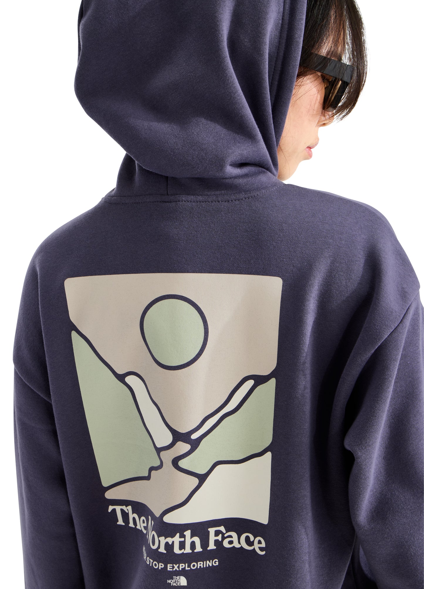 A person wears a dark blue hoodie featuring a back graphic with a stylized nature scene of rolling hills, a winding river, and a sun, accompanied by the text The North Face and Never Stop Exploring.