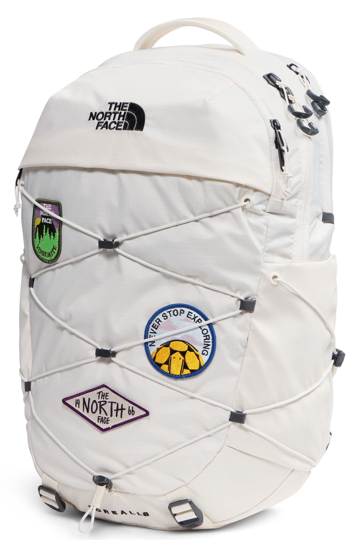 A cream-colored backpack with a bungee cord system on the front, decorated with several circular and diamond-shaped patches featuring graphics and text including 'NEVER STOP EXPLORING' and 'THE NORTH FACE 66'.