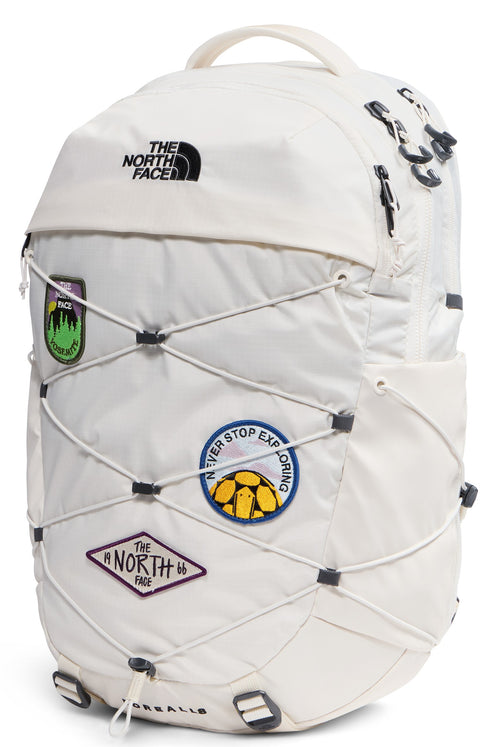 A cream-colored backpack with a bungee cord system on the front, decorated with several circular and diamond-shaped patches featuring graphics and text including 'NEVER STOP EXPLORING' and 'THE NORTH FACE 66'.