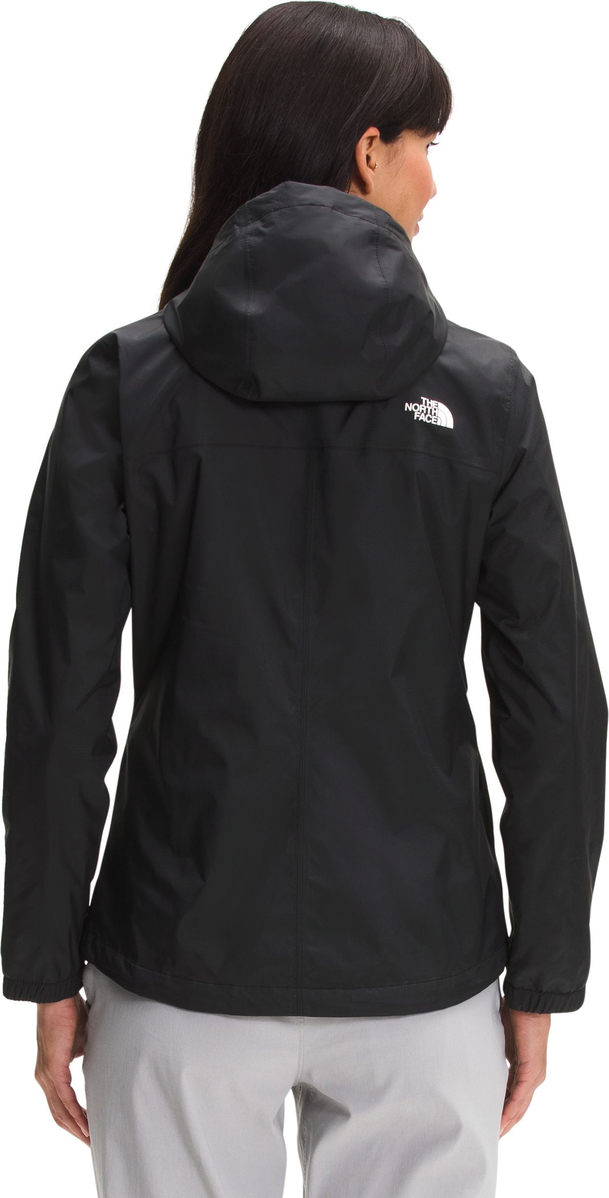 A back view of a woman wearing a black, long-sleeved hooded rain jacket with a white logo on the upper right back, paired with light gray pants.