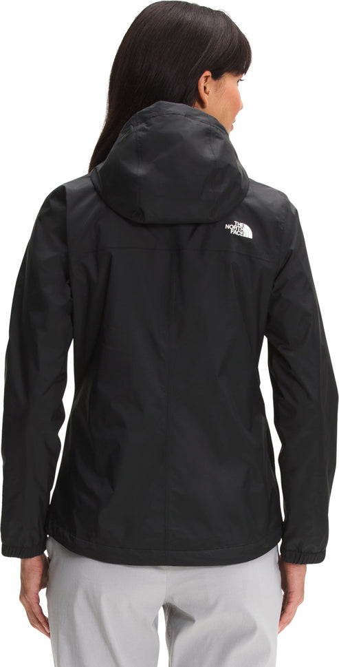 A back view of a woman wearing a black, long-sleeved hooded rain jacket with a white logo on the upper right back, paired with light gray pants.