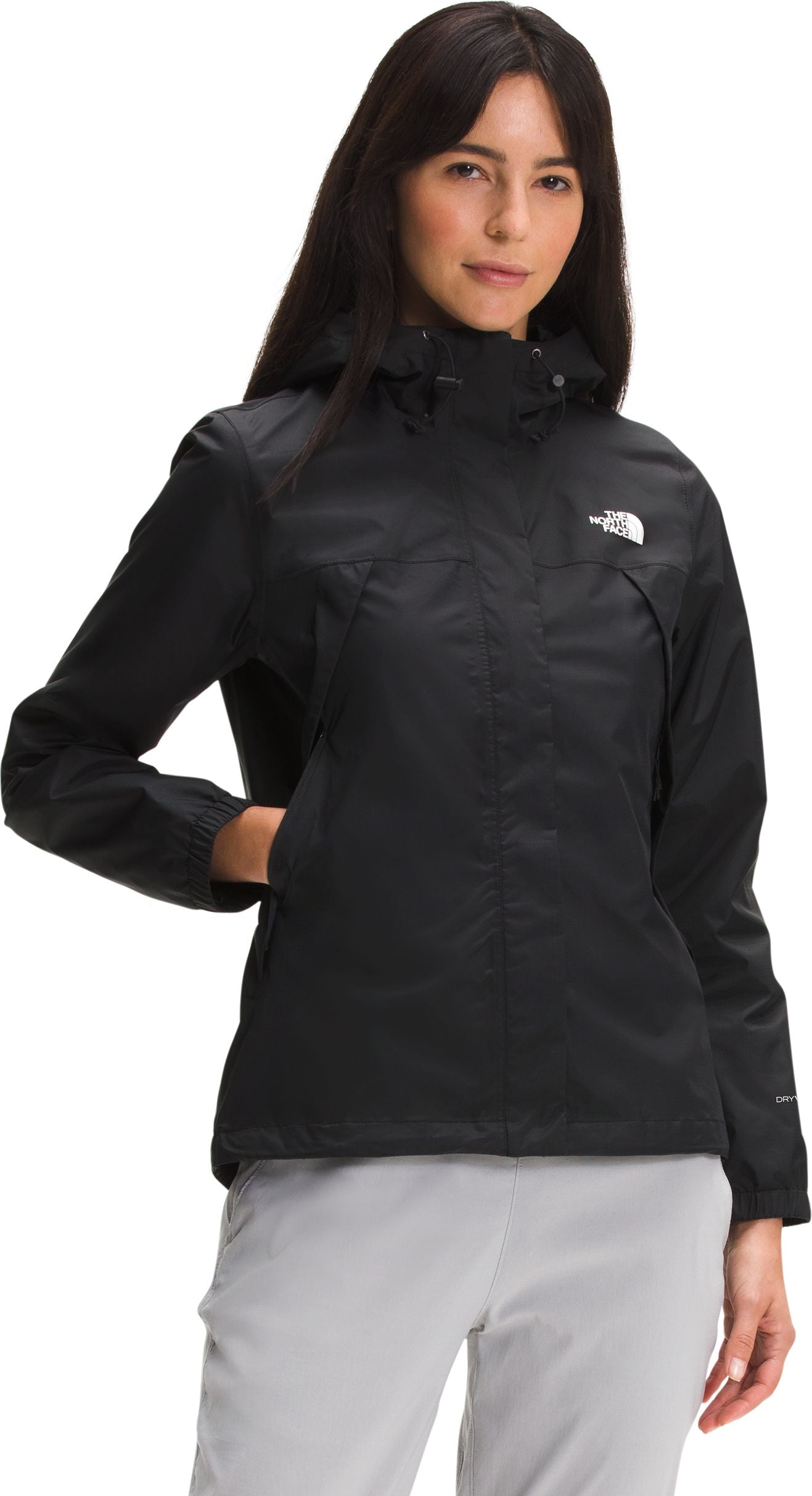 A woman wearing a black, hooded, long-sleeved windbreaker jacket with a zipper closure and a white logo on the upper left chest, paired with light gray pants.