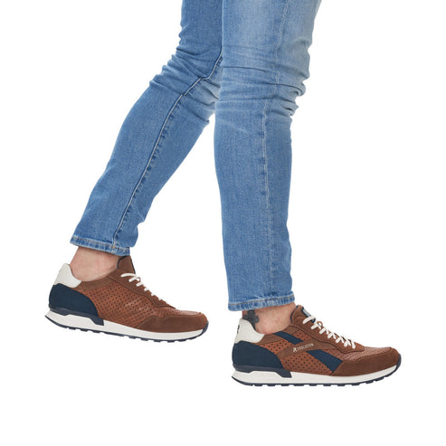 A person wearing jeans and perforated brown sneakers with white soles and blue accents.