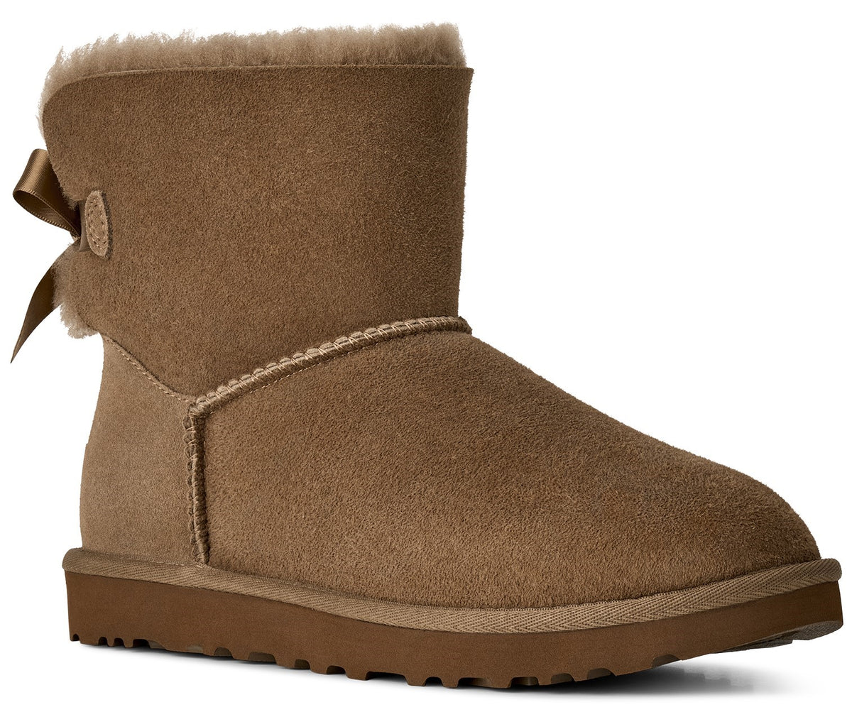 A tan suede ankle boot featuring a thick faux fur lining, a solid brown rubber sole, and a decorative ribbon bow attached to the outer side.