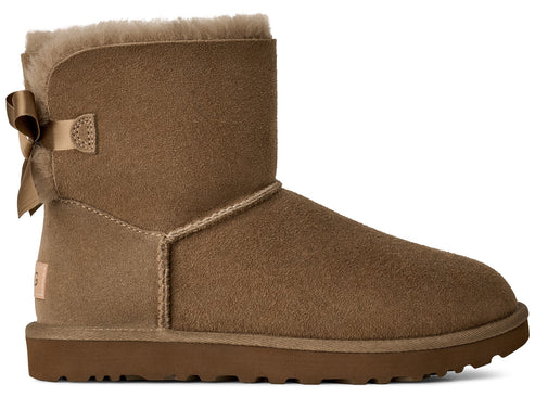 A tan suede ankle boot featuring a fluffy wool interior, a satin ribbon bow on the heel, and a thick brown rubber sole.