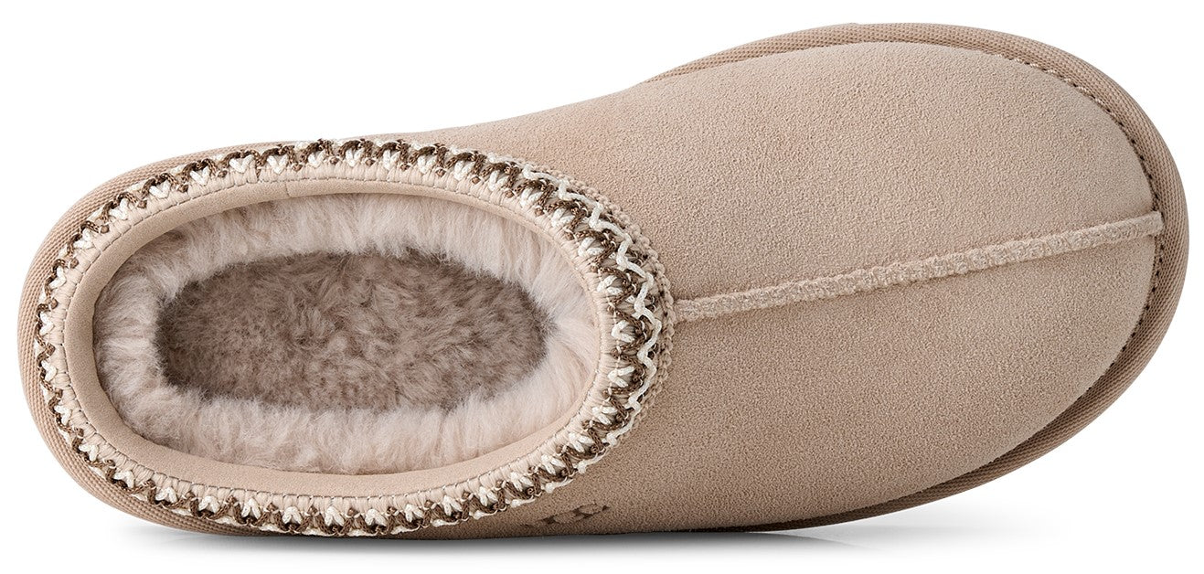 A tan suede slip-on slipper with a fluffy faux-fur lining and a decorative braided trim around the opening.