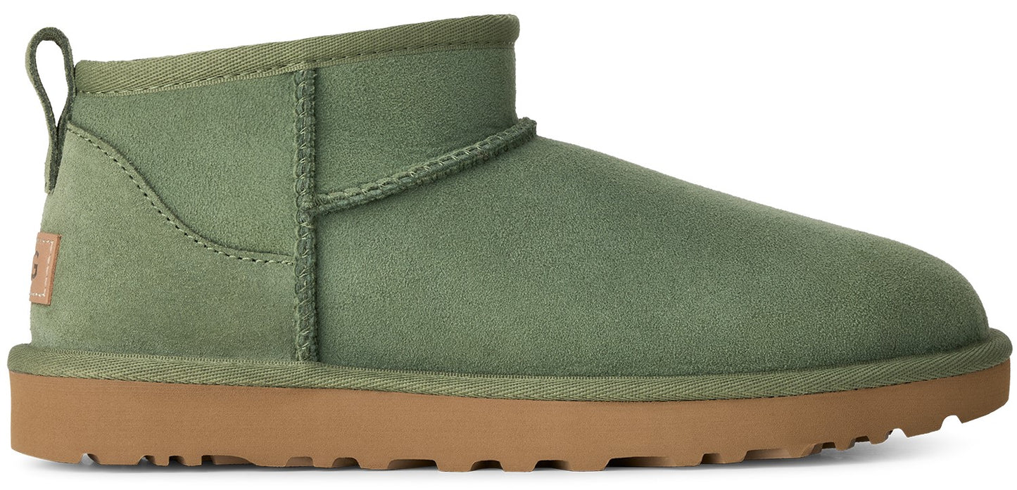 A sage green suede ankle boot with a pull loop on the back and a flat, tan rubber sole, showing a small tan tag on the heel.