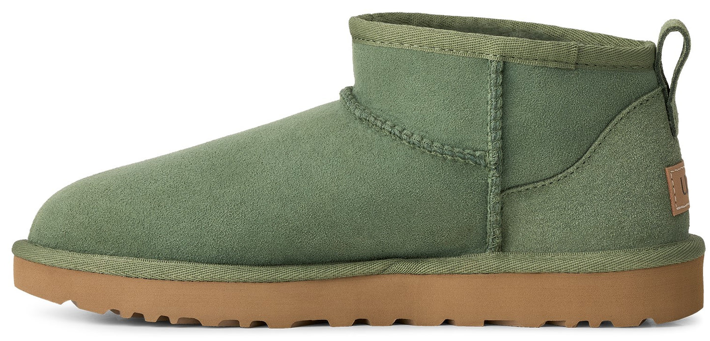 A sage green suede ankle boot featuring a tan gum rubber sole and a small rectangular logo tag on the heel.