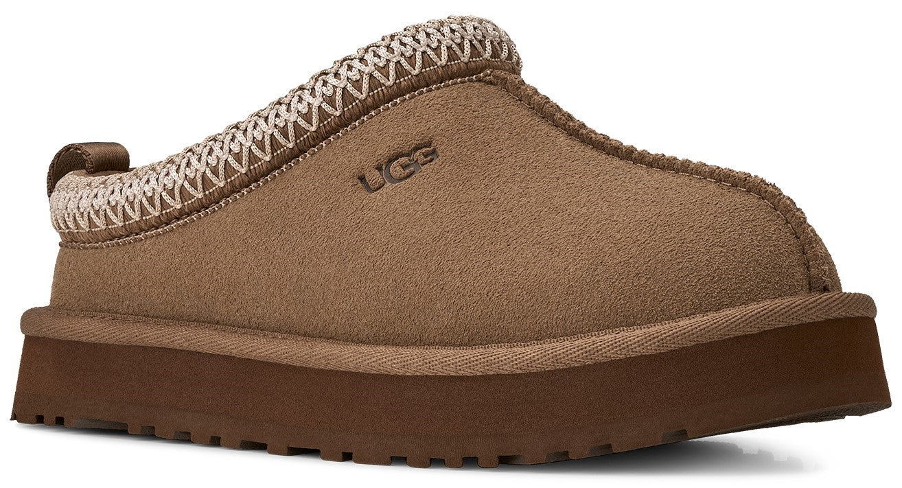 A tan suede platform slipper with a thick brown rubber sole, a woven beige and brown trim along the collar, and the logo UGG embossed on the side.