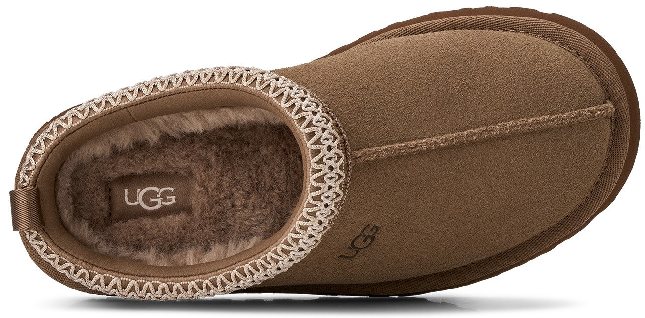 A tan suede slipper seen from above, featuring a decorative woven cream-colored border around the opening, a soft brown fur-lined interior with the UGG logo visible on the insole, and a small UGG logo embossed on the top of the upper.