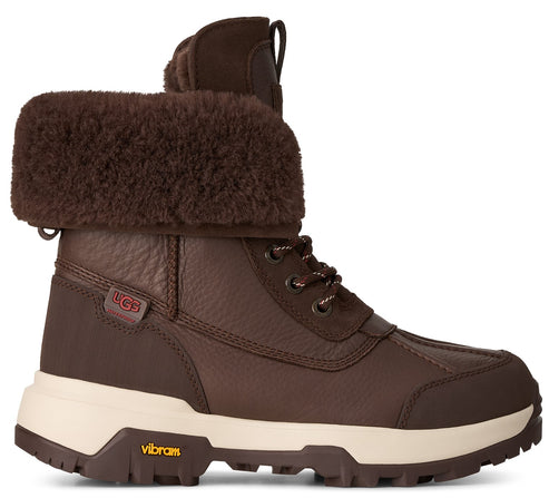 A brown leather UGG winter boot with a thick, fold-over faux fur collar, white midsole, and Vibram rubber outsole, viewed from the side.