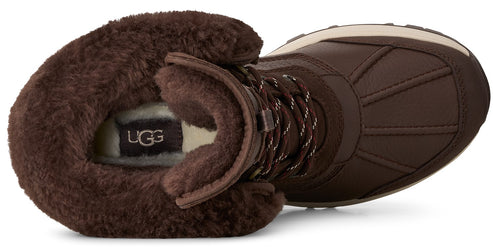 A brown winter boot shown from a top-down angle, featuring a thick, fuzzy brown fleece lining around the opening, a visible UGG brand patch inside, and a brown leather outer shell with a reinforced, stitched toe box.