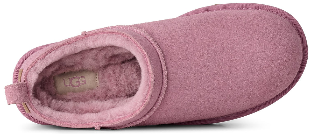 A top-down view of a light pink, suede slip-on shoe with a thick, plush, fuzzy interior lining and an UGG logo patch on the insole.