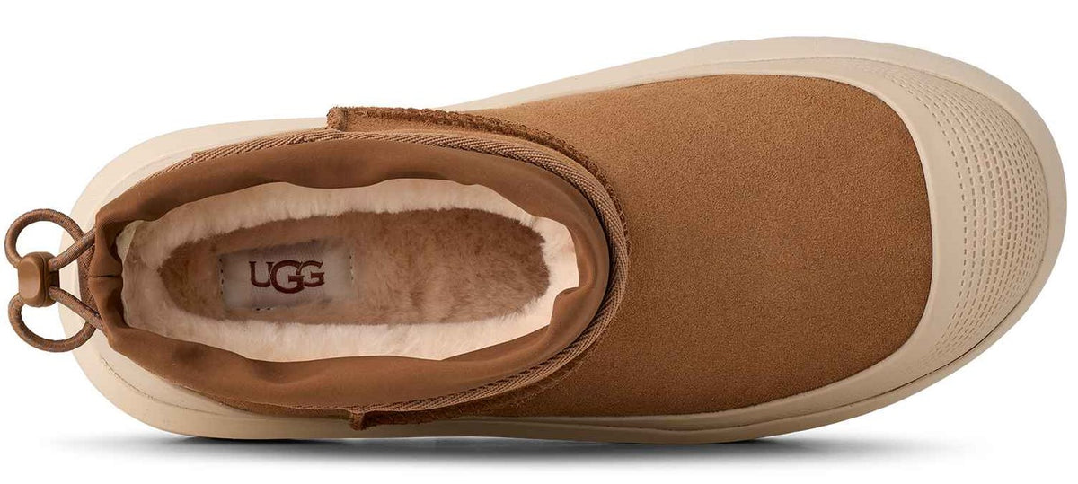 A top-down view of a tan suede slip-on shoe with a cream-colored fleece lining and an UGG logo patch on the insole, featuring a rounded toe and a cinch cord at the heel.