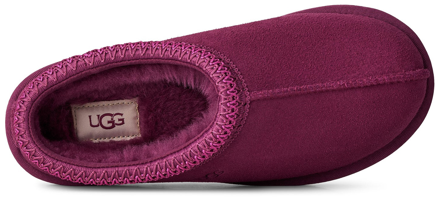 A top-down view of a dark magenta suede slipper featuring a soft, fuzzy interior lining and a decorative braided trim around the opening, with an UGG label visible inside.