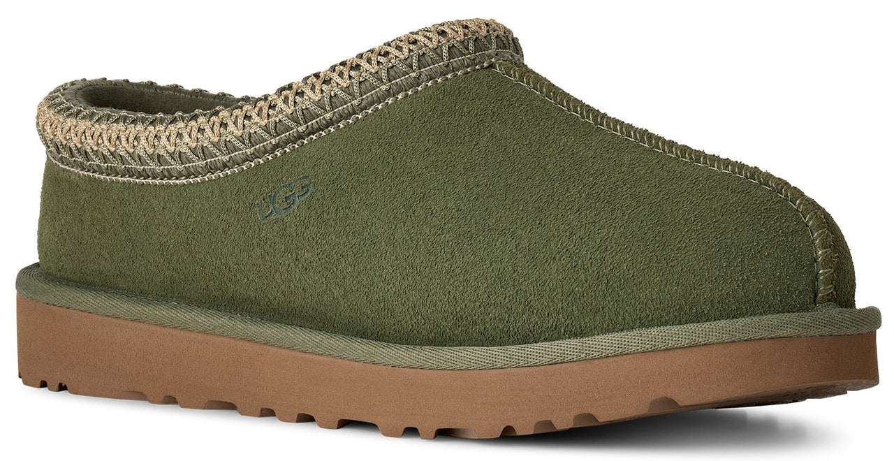 A sage green suede slipper with a woven beige trim around the collar, a rounded toe, and a flat, light brown rubber sole.