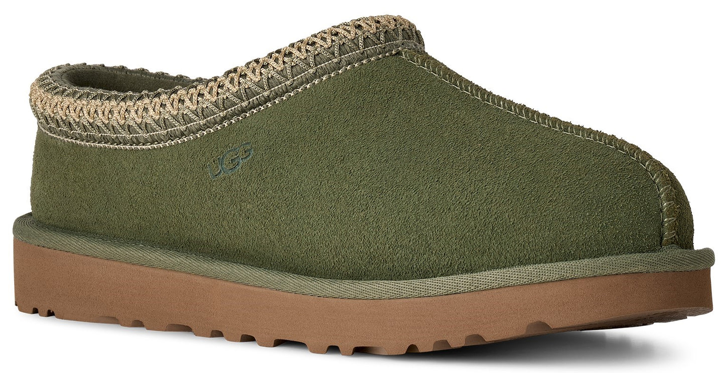 A sage green suede slipper with a woven beige trim around the collar, a rounded toe, and a flat, light brown rubber sole.