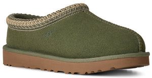 A sage green suede slipper with a woven beige trim around the collar, a rounded toe, and a flat, light brown rubber sole.