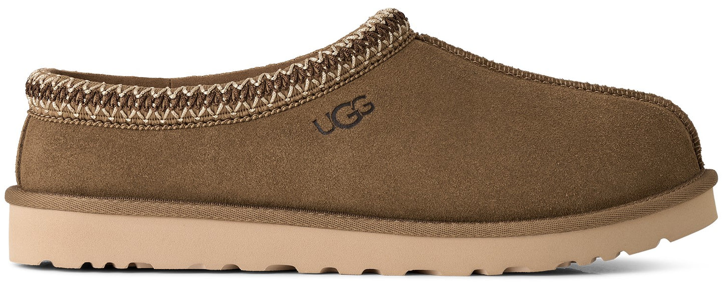 A brown suede slip-on shoe with a cream-colored sole and a woven decorative trim along the collar, featuring the text UGG embossed on the side.