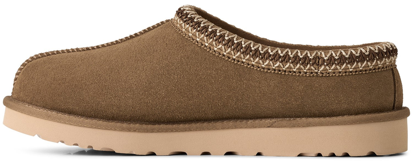 A side view of a tan suede slip-on slipper with a thick, flat light beige rubber sole and a decorative braided trim around the collar.